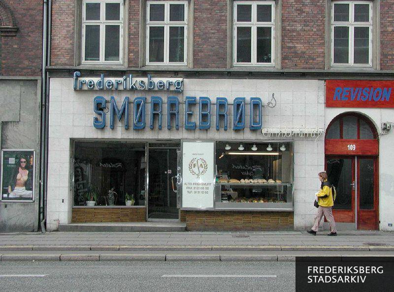 Butiksfacade til Frederiksberg Smørrebrødsanretning, ved siden af ses en rød dør og indgangen til en anden virksomhed skiltet med Revisor
