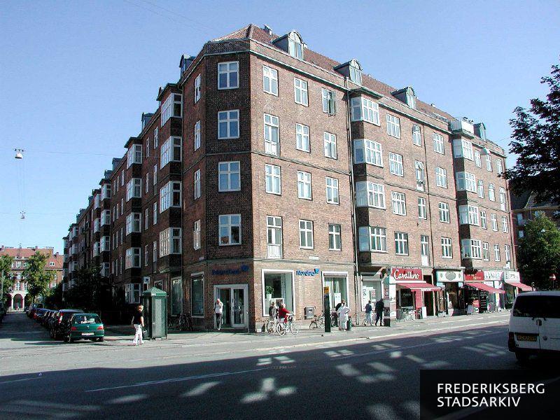 Hjørneejendommen Peter Bangs Vej 60 med butiksfacader til banken Nordea, sandwich-baren Cavallino, en dametøjsbutik, cykelsmeden og cykelhandleren Cykler og Emil Slomanns Vej 2, hvor Lindevangsskolen skimtes for enden af vejen bag træerne denne solskinsdag med blå himmel. Der er fodgængere og cyklister, der trækker deres cykler. Desuden ses en gammel reklamesøjle med kobbertag, et busstoppested uden læskur, en telefonboks og parkerede biler. Lyset fra de sprossede vinduer er genspejlet på den del af vejbanen, der ligger i skygge