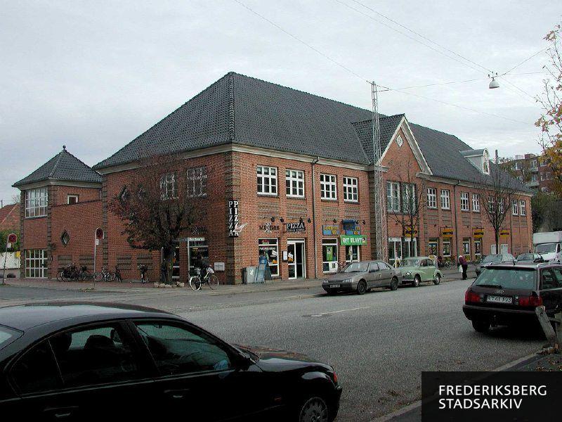 Hjørneejendommen Peter Bangs Vej 100-104 og Gustav Johannsens Vej 2A-G med butiksfacader til Maximus Pizza, Blockbuster Video og Netto