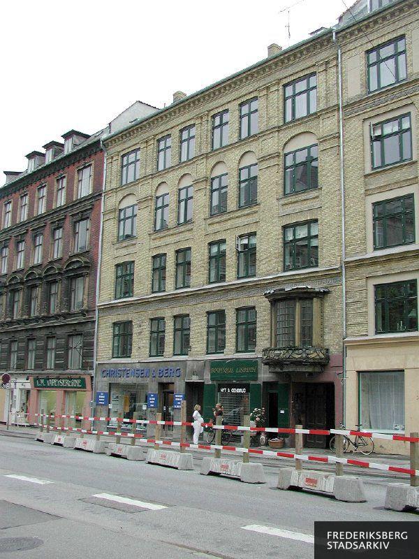 Ejendom og butiksfacader til Blikkenslager VVS H.P. Christensen & Chr. Berg A/S og genbrugsbutikken Boutique Lisbeth med afspærring til vejarbejde foran. To yngre kvinder taler sammen på fortovet. I naboejendommen ses butiksfacaden til L.M. Tapetsererne