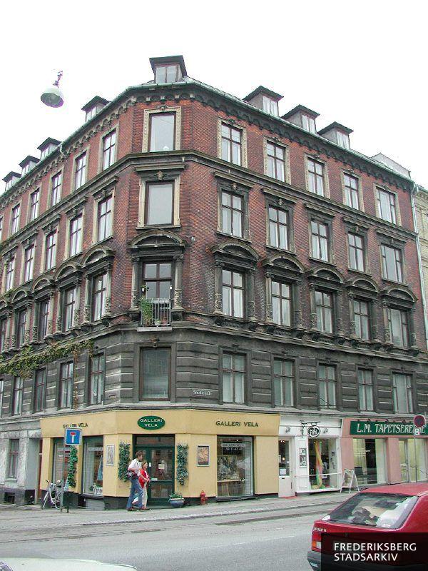 Hjørneejendommen Smallegade 43 og Virginiavej 2 med butiksfacader til Galleri Vela på hjørnet, Salon Ami med A-skilt på fortovet og til højre L.M. Tapetsererne. Der er lidt bevoksning, måske grøn vin, på facaden på Virginiavej. Tre vinduer på det afskårne hjørne er usprossede som butiksvinduerne