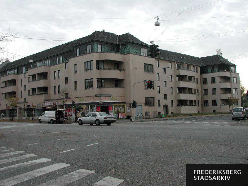Hjørneejendommen Jens Jessens Vej 1-3 og Peter Bangs Vej 103-107 med butikker, kiosk, Kina-Grill, lyskryds i forgrunden, bilister, cyklister og en åben varevogn