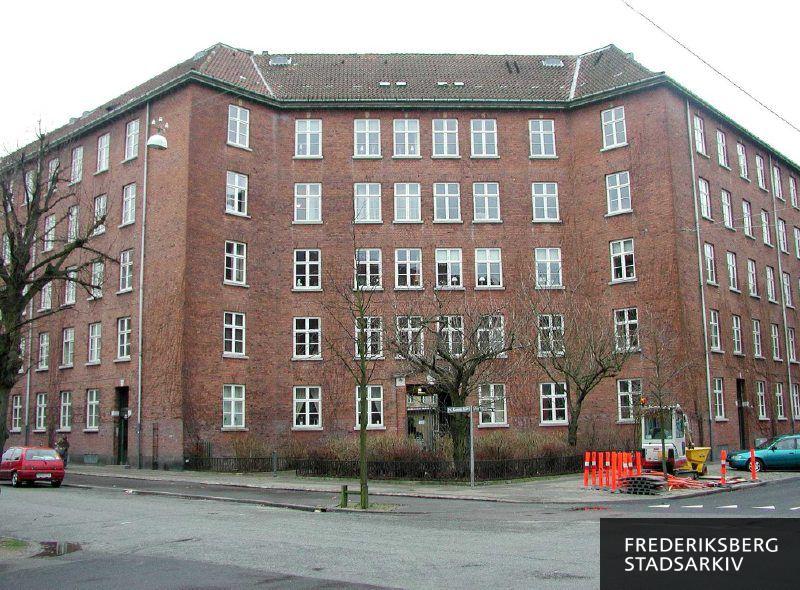 Det markante, afskårne hushjørne med indhegnede træer og buske ved Julius Valentiners Vej 29 og P.G. Ramms Allé 6. På fortovet står materialer, udstyr og redskaber til vejarbejde