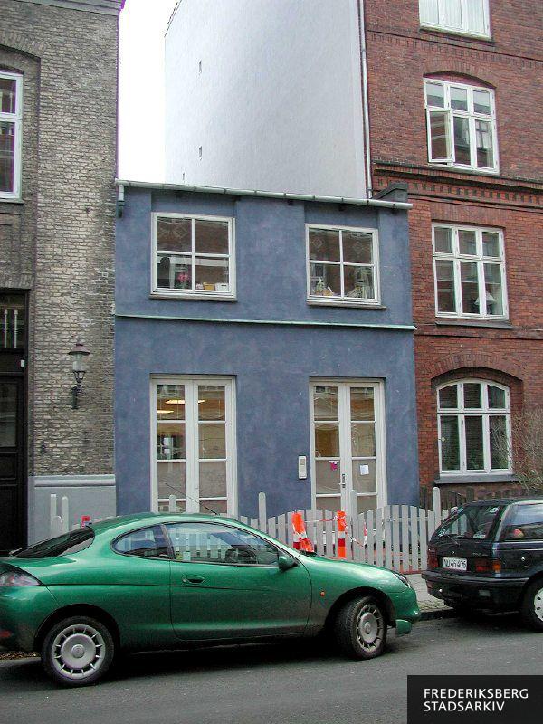 Den blå to-etages ejendom med hvidt stakit mellem højere etageejendomme i 2006, i nr. 8A findes dagplejeordningen "Min lille verden". Et farverigt foto med et gult, et blåt og et rødt hus, en hvid gavl, orange afspærring foruden en grøn og en sort bil parkeret i forgrunden. Ved naboejendommens gadedør er der en gammel lampe på facaden