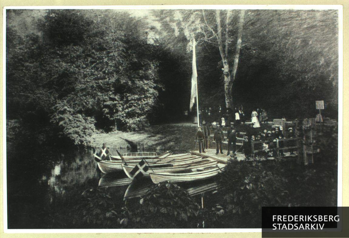 Den første bådebro for Frederiksberg Haves bådfart i kanalerne. Den første bådejer Chr. Svendsen (1835-1907) - den dekorerede mand - står midt på broen. Drengen ved flagstangen er hans søn, den anden bådejer Chr. Svendsen (1869-1922)