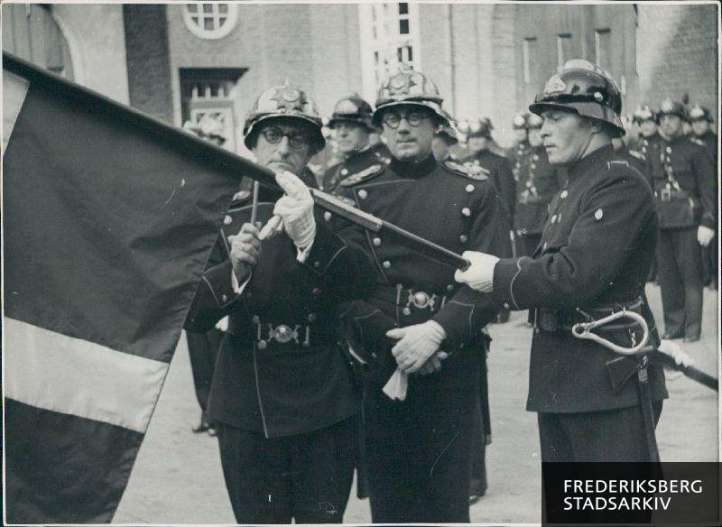 En ny korpsfane indvies i forbindelse med brandvæsenets 75 års-jubilæum. 
Brandchef Bang slår andet søm i (efter borgmesteren slog første søm i).
Fra venstre mod højre ses udover Bang: Brandingeniør J. S. Eilertsen og nr. 94 Erik Velin