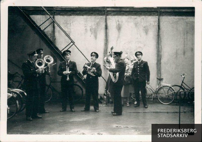 Musikforeningen spiller i anledning af sølvbryllup. Musikanterne i uniform med instrumenter, horn, fra venstre: Liltorp, Zimling, Handryciak, Raith, V. K. Jensen og E. H. Olsen længst til højre. I baggrunden står nogle damer og vinker, måske til dagens ægtepar.