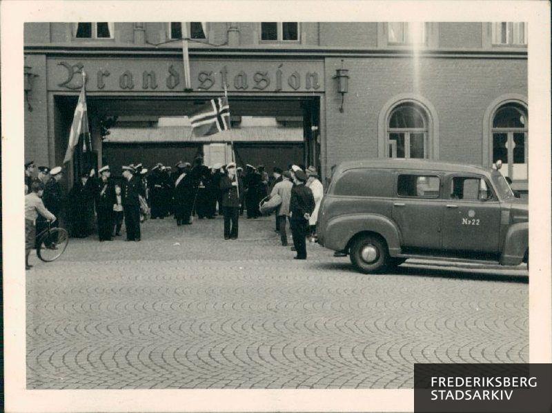 Der lægges an til parade gennem byen, Bergen, med musikforeningen fra Frederiksberg Brandvæsens deltagelse, i anledning af den norske nationaldag. Billedet er fra indgangsporten til Bergens brandstation på Skansen og både dannebrog og det norske flag er foldet ud.