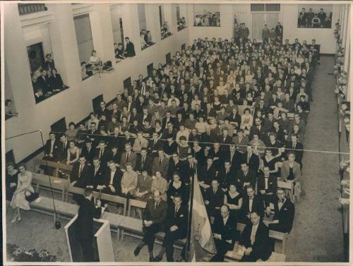 Skolens hvor en mængde personer er Andre personer står i nicherne. Talerstol med mikrofon. Skolens 25 års jubilæum fejres (januar 1959) kbhbilleder.dk