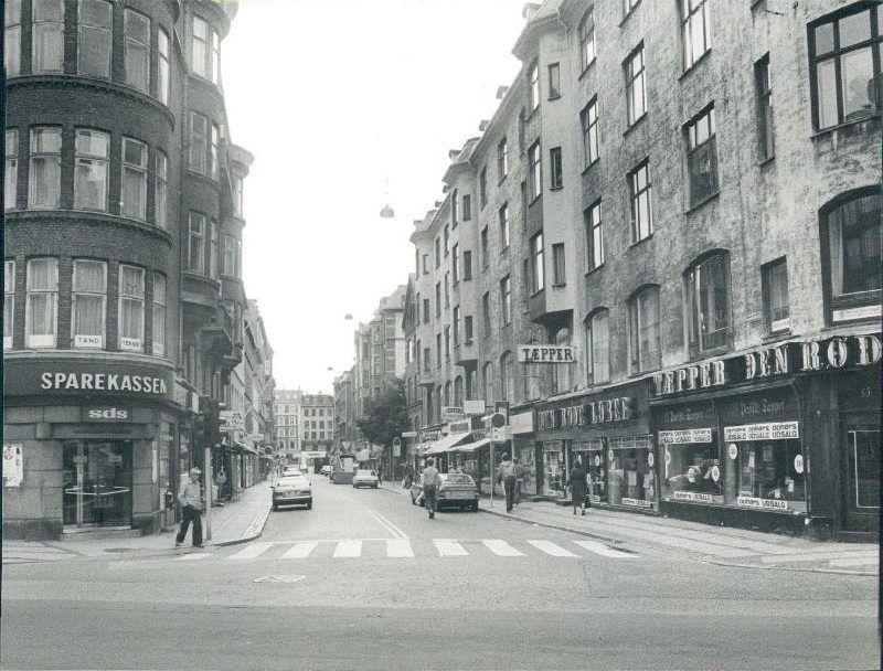 Værnedamsvej set fra Gammel Kongevej ned mod Vesterbrogade/Frederiksberg Allé. Til venstre i den store fireetages beboelsesejendom med buede karnapper ses en afdeling af sparekassen SDS, og på det højre hjørne ligger tæppeforretningen "Den røde løber". Der er i begge sider parkerede biler, og der ses fodgængere på både kørebane og fortove
