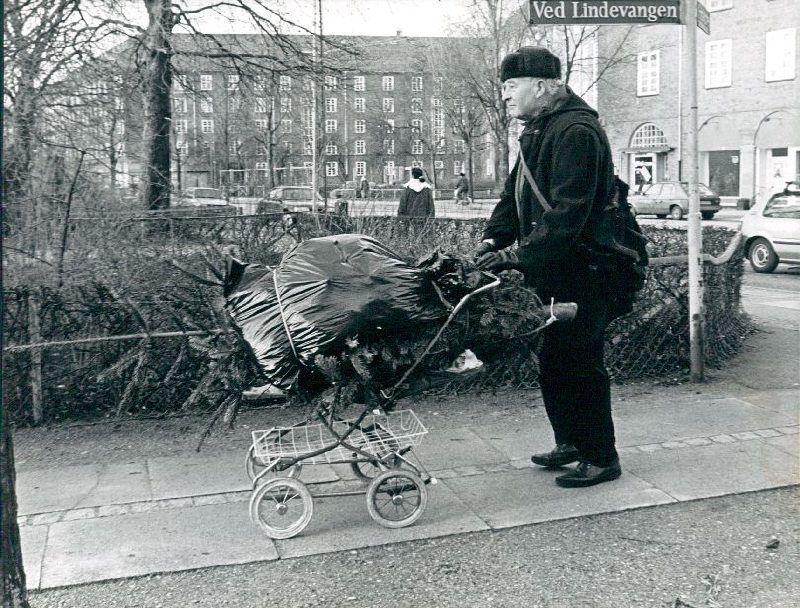 En varmt påklædt herre transporterer på en lille firehjulet vogn et grantræ, som er emballeret i plastik
Fotoet er taget på hjørnet af Ved Lindevangen og Finsensvej
I baggrunden ses etagehuse på Dalgas Boulevard og til højre for personen i billedet ses hjørnet af Lindevangsparken