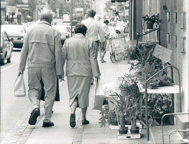 Godthåbsvej - et ældre par spadserer med hinanden i hånden. Der er parkerede biler, og til højre i billedet ses udstilling af blomster