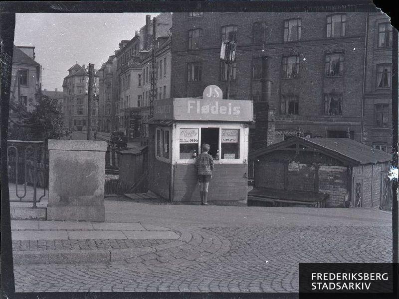 Isbod, "JS Flødeis" på Nordre Fasanvej. Adressen på ejendommen bag ishuset er Howitzvej 71. I nærheden af denne anes jernbanelinjen, Frederiksberg-Vanløse-banen, med tilhørende bomhus.