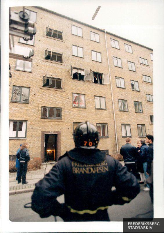 Et tv brød i brand i en lejlighed på Ane Katrinesvej 28, En brandmand står på gaden og kigger op mod lejligheden, der har vidt åbne vinduer. Politifolk taler med tilskuere og beboere på fortorvet