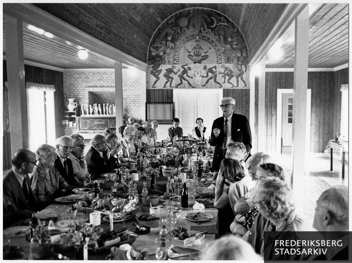 Emil Velmar holder tale ved frokostbordet for pensionister fra Frederiksberg Kommunelærerforening på den årlige skovtur. I baggrunden ses kommunelærer Jørgen Thoms vægdekoration