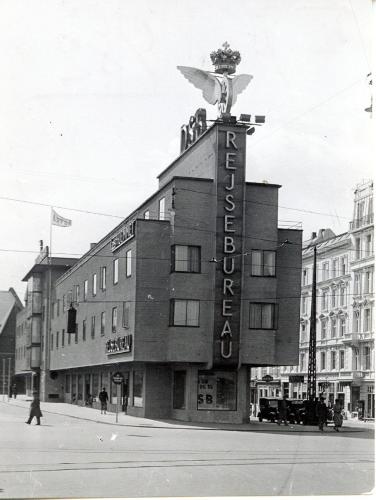 Københavns 3. Hovedbanegård: dsb-logo, Vingehjul, flag, flagstang, facadereklame, lyskurv, fodgænger, sporvognsskinner, bil, personbil, fortov, kantsten, Reventlowsgade, Vesterbrogade, fordakning, gesims, gittermast, lygtepæl, trafikskilt, Hotel Astoria, taxa, taxaholdeplads